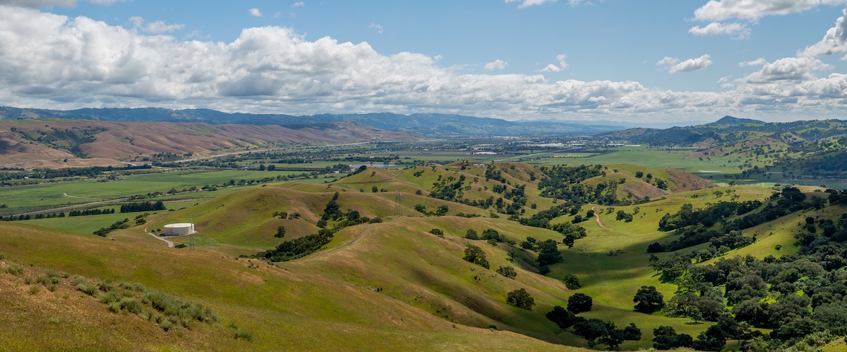 Expansive view looking south across Coyote Valley, with green fields, hillsides spotted with trees, mountains extending into the distance, and a sky filled with white fluffy clouds
