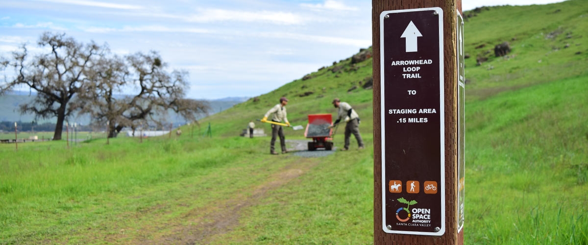 Arrowhead Loop trail marker in foreground, out of focus in the background are two Open Space Technicians in tan uniforms shoveling gravel onto the trail