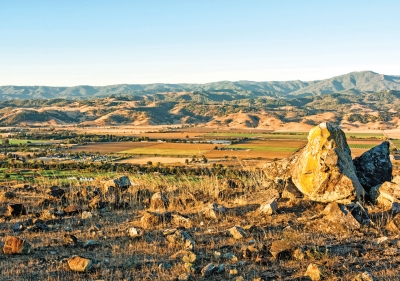 Looking down from a rock-covered hilltop at an expansive view of Coyote Valley agricultural fields, the Santa Cruz foothills and mountains in the distance