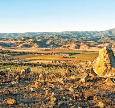 Looking down from a rock-covered hilltop at an expansive view of Coyote Valley agricultural fields, the Santa Cruz foothills and mountains in the distance