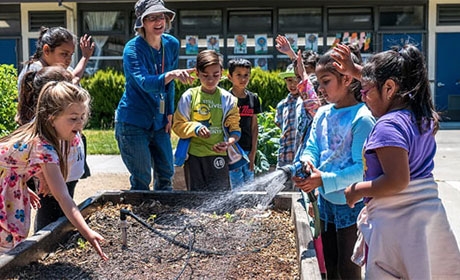 Smiling students and teacher around raised garden bed with sprinkler hose