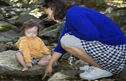 Mother in blue shirt and checkered skirt pointing something out to toddler in orange shirt sitting on rocks
