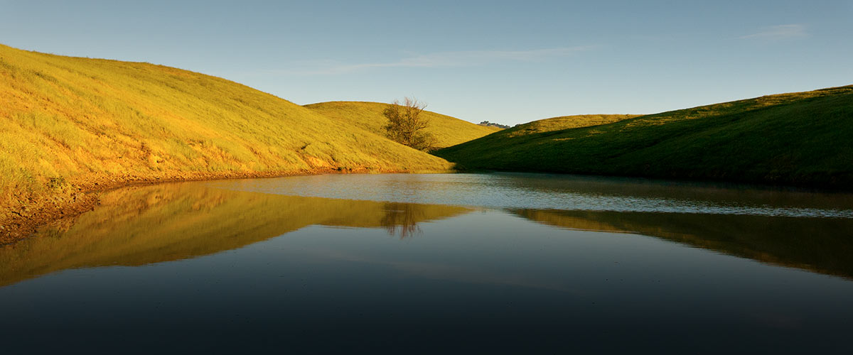 Full pond surrounded by bright green hills under a clear blue sky, the water reflecting the hillsides and sky