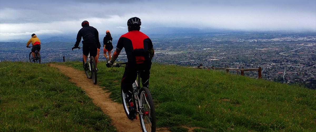 Three mountain bikers and two hikers traveling along dirt trail at top of grassy hill overlooking urban San Jose below